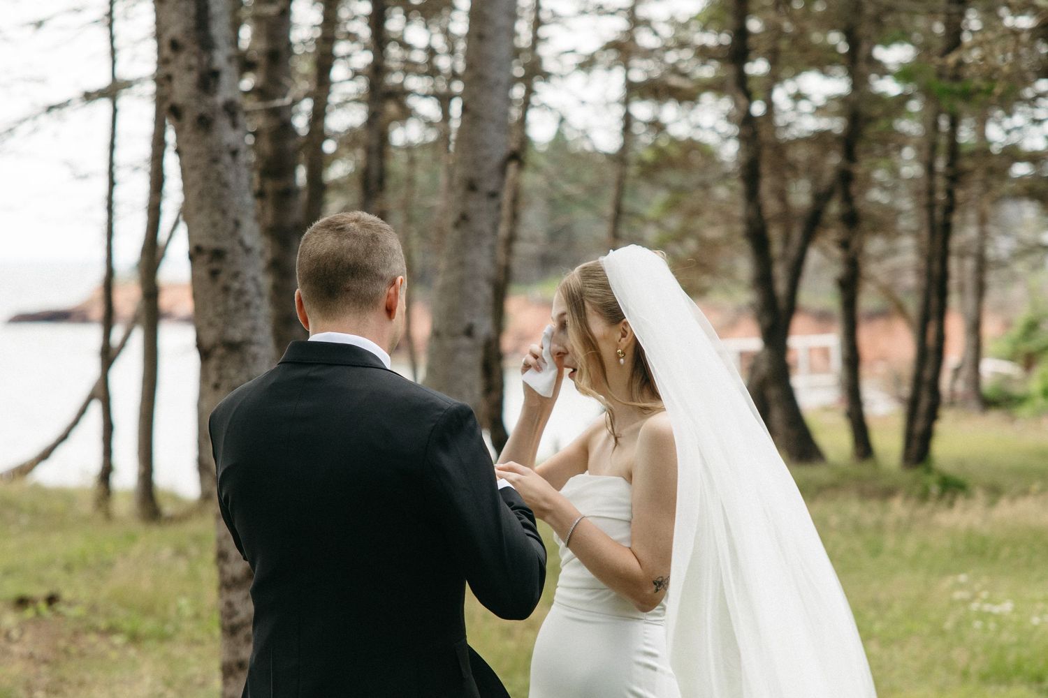 Bride in white gown and veil exchanging vows with groom in black suit outdoors near pine trees.