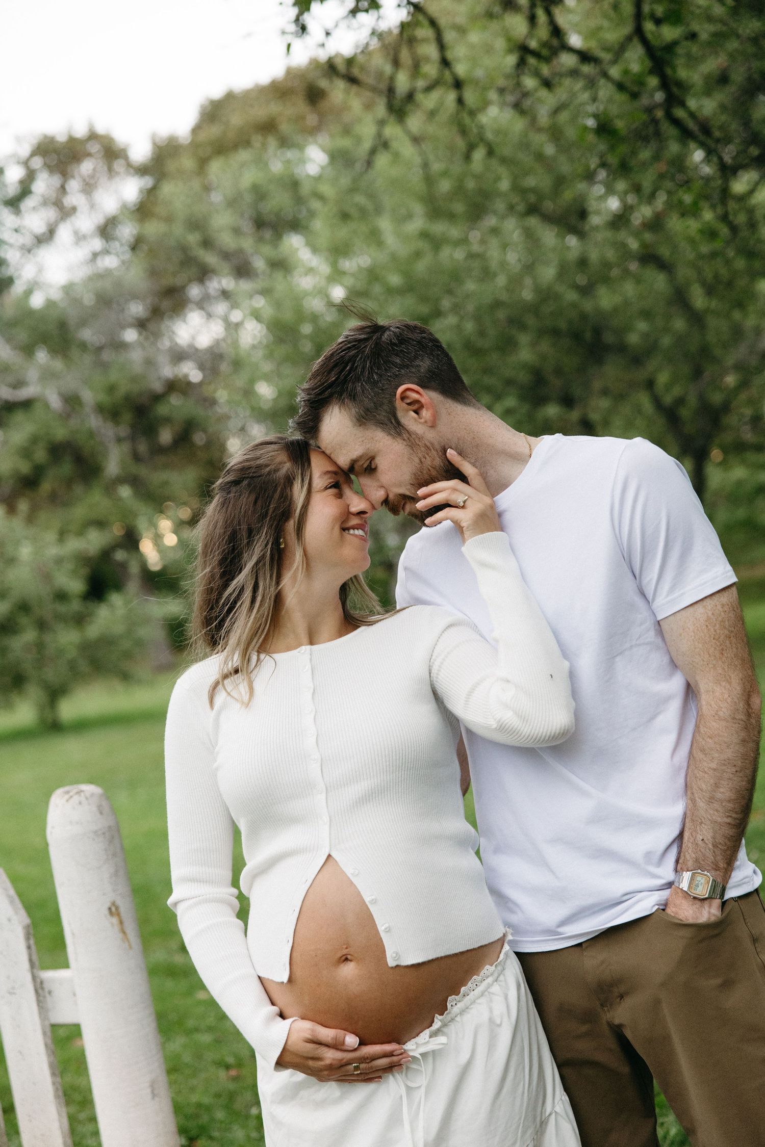 Pregnant couple in white outfits sharing a tender moment outdoors, woman cradling baby bump near white fence.