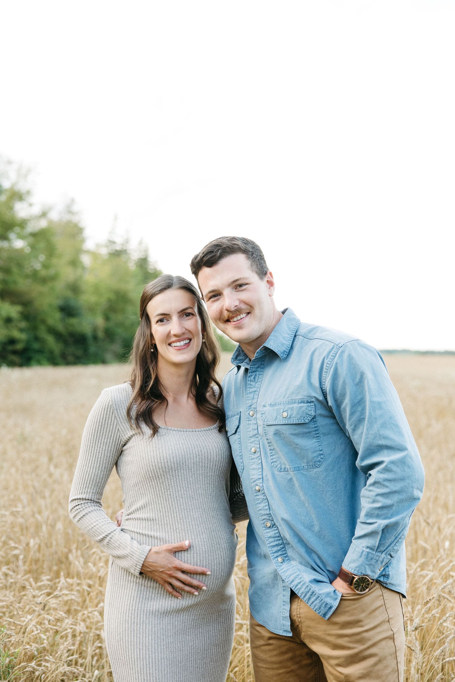 Smiling pregnant woman in gray dress and man in denim shirt pose together in wheat field.