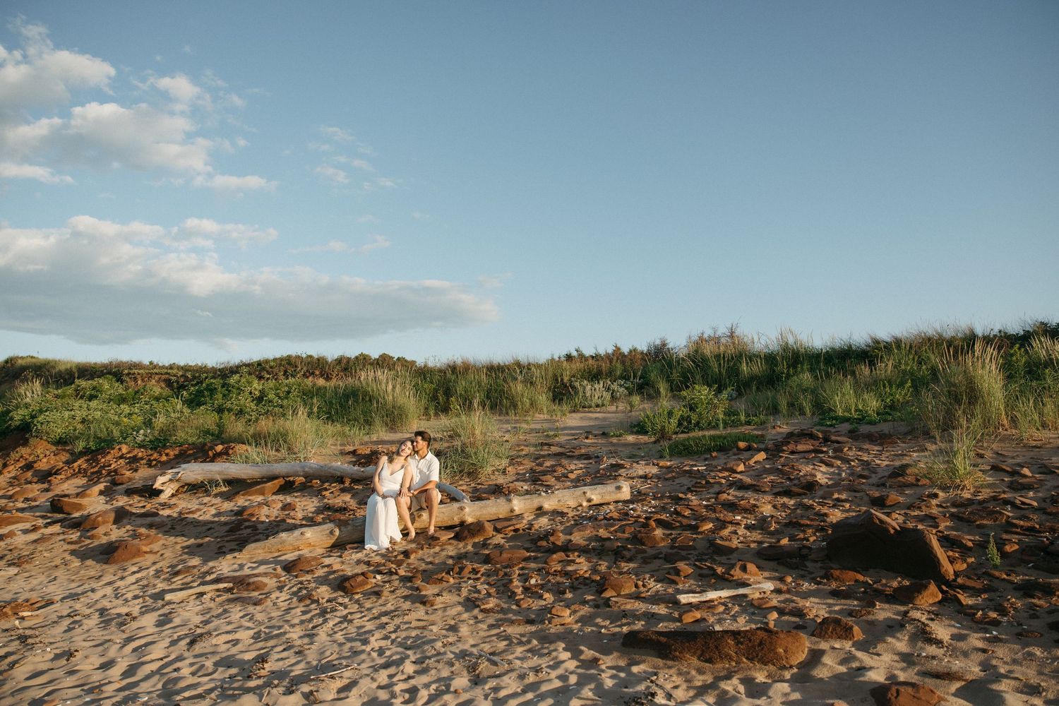 Couple sitting together on driftwood logs on a sandy beach with dunes and greenery behind them.