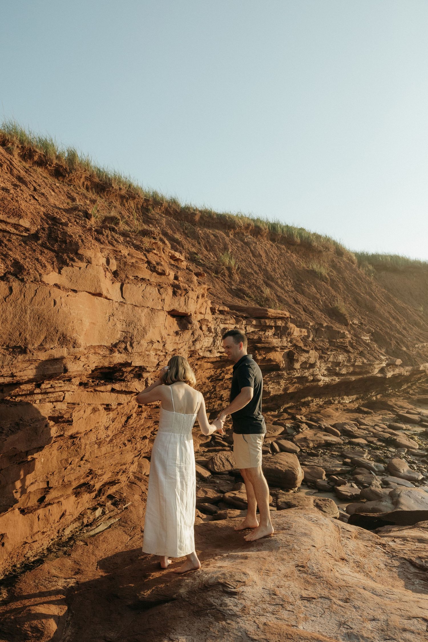 A couple stands on rocky coastal cliffs with red sandstone formations.