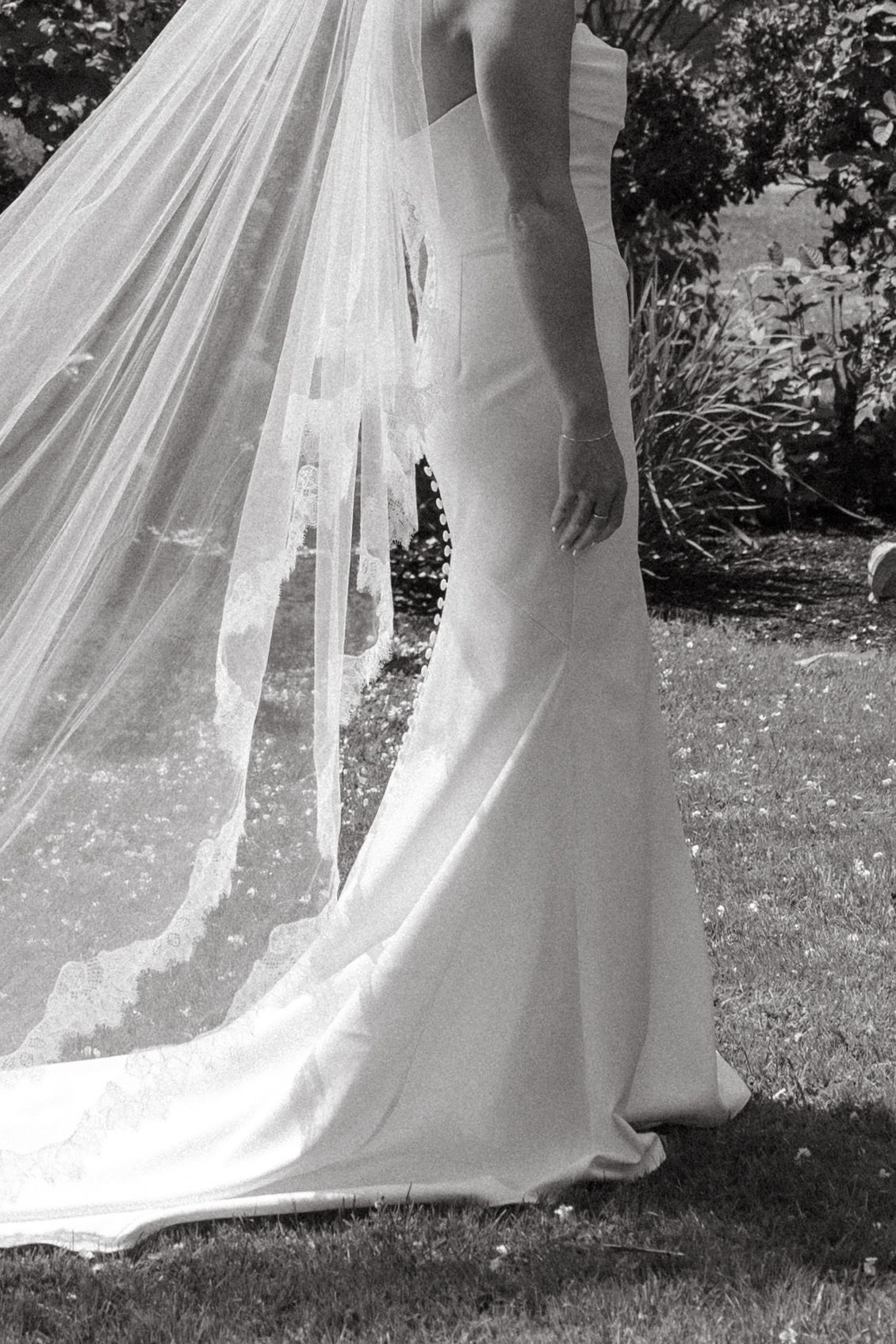 Close-up details of a flowing white wedding dress and veil against green grass.