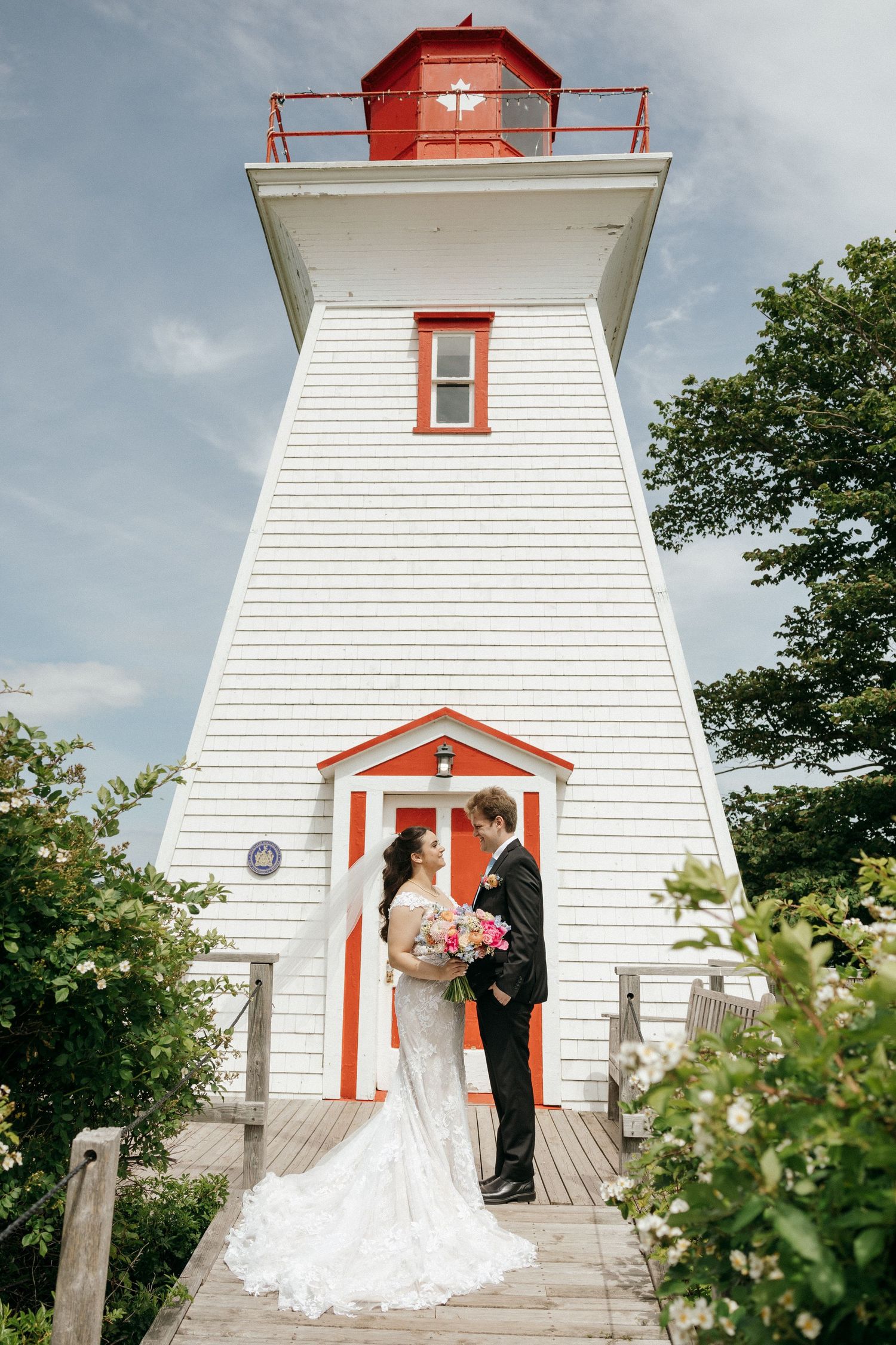 A white lighthouse with red trim frames a romantic wedding scene on its wooden deck.