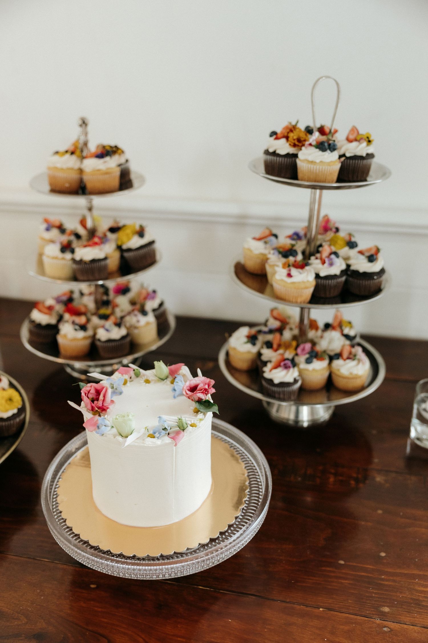 Wedding dessert display featuring tiered cake stands with cupcakes and decorated white cake.