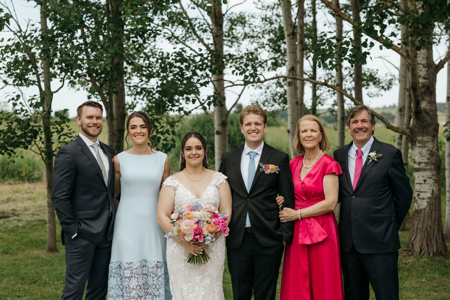 Wedding party in formal attire posing outdoors with trees in background.