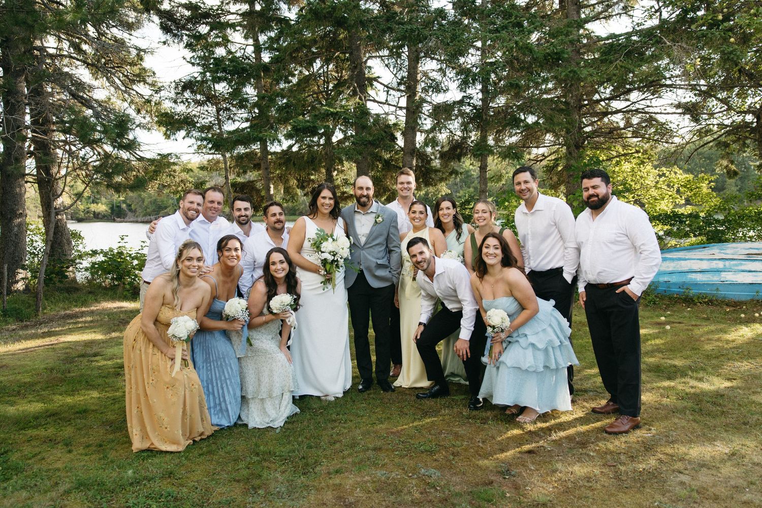 Wedding party in light blue and white attire poses together outdoors under tall evergreen trees.