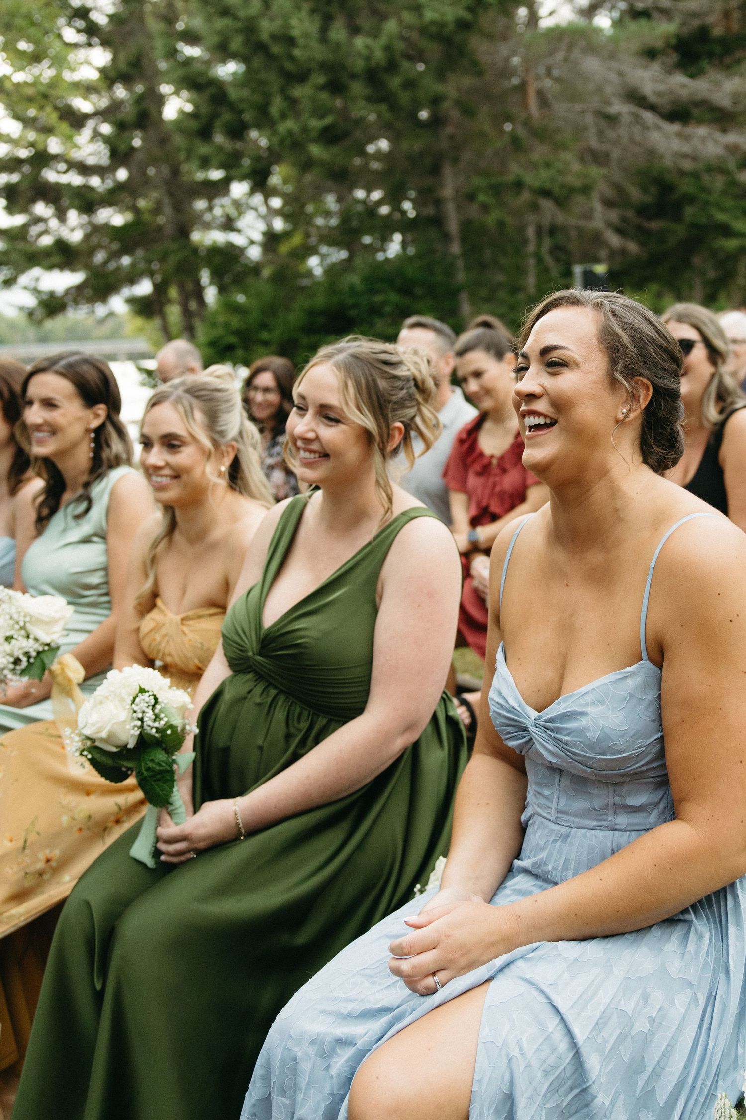 Wedding party wearing blue and green dresses holding white bouquets while seated outdoors.