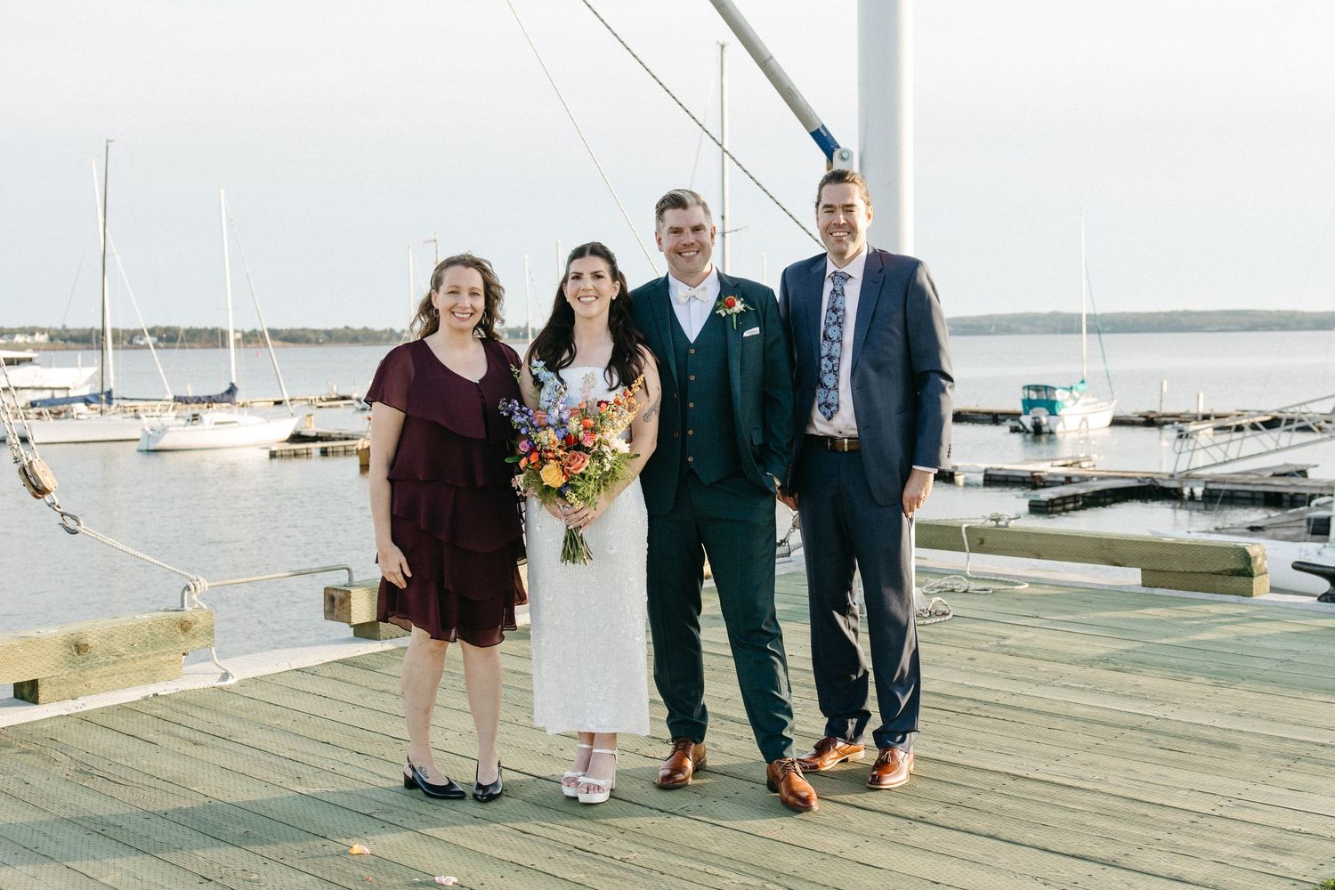Bride holding a colorful bouquet poses with three wedding guests on a wooden dock overlooking sailboats and calm harbor waters.
