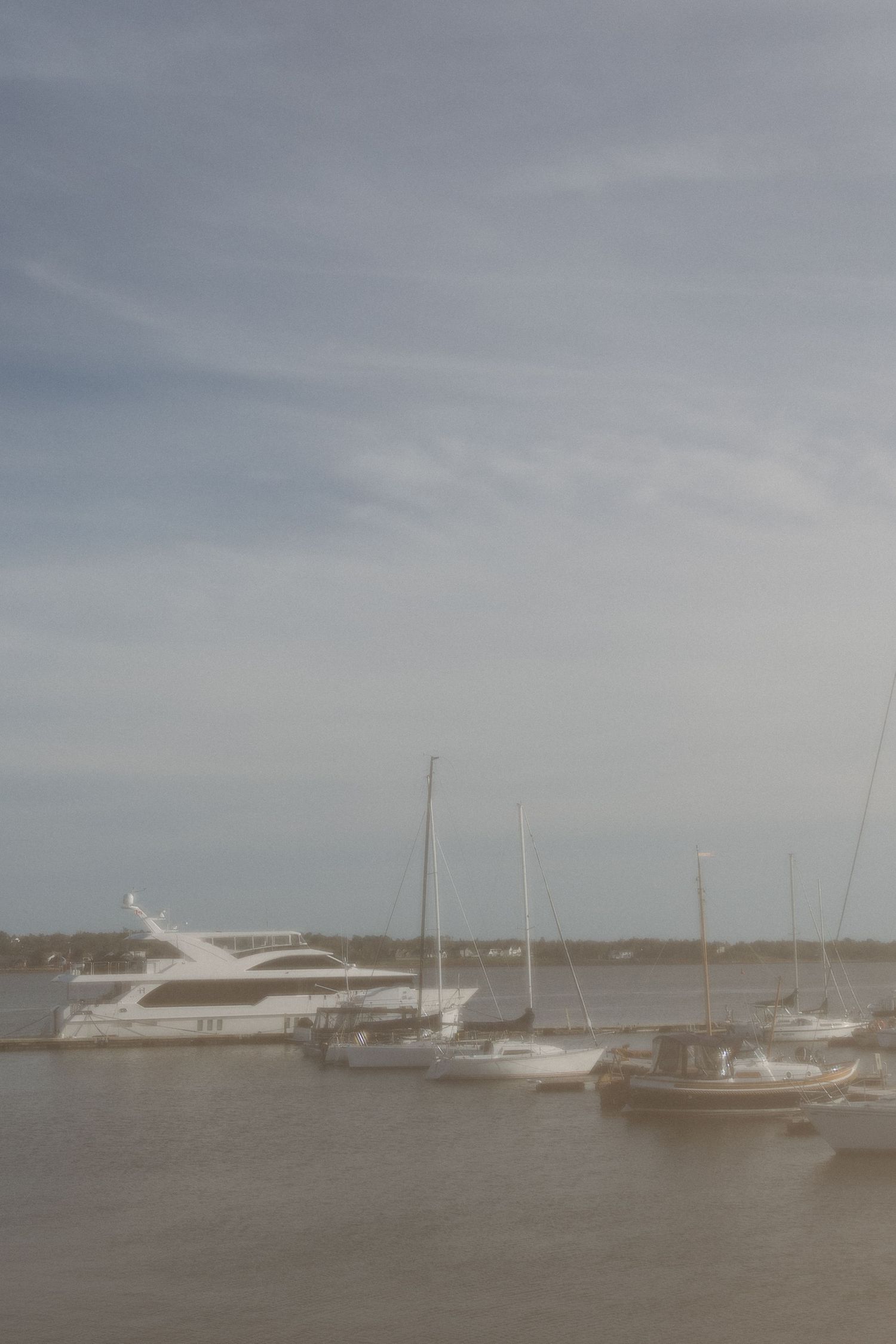 White motor yacht and sailboats moored in a calm harbor under a soft, hazy, overcast sky.
