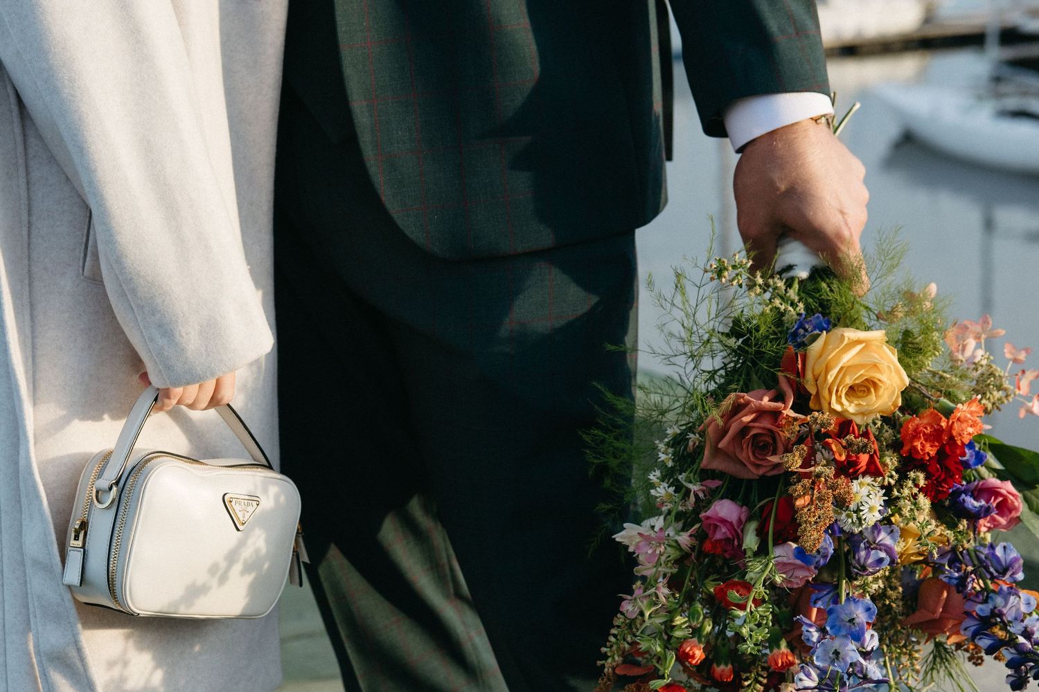 Groom in a dark suit holds a colorful bouquet of roses and wildflowers beside a bride in white with a small handbag near the water.