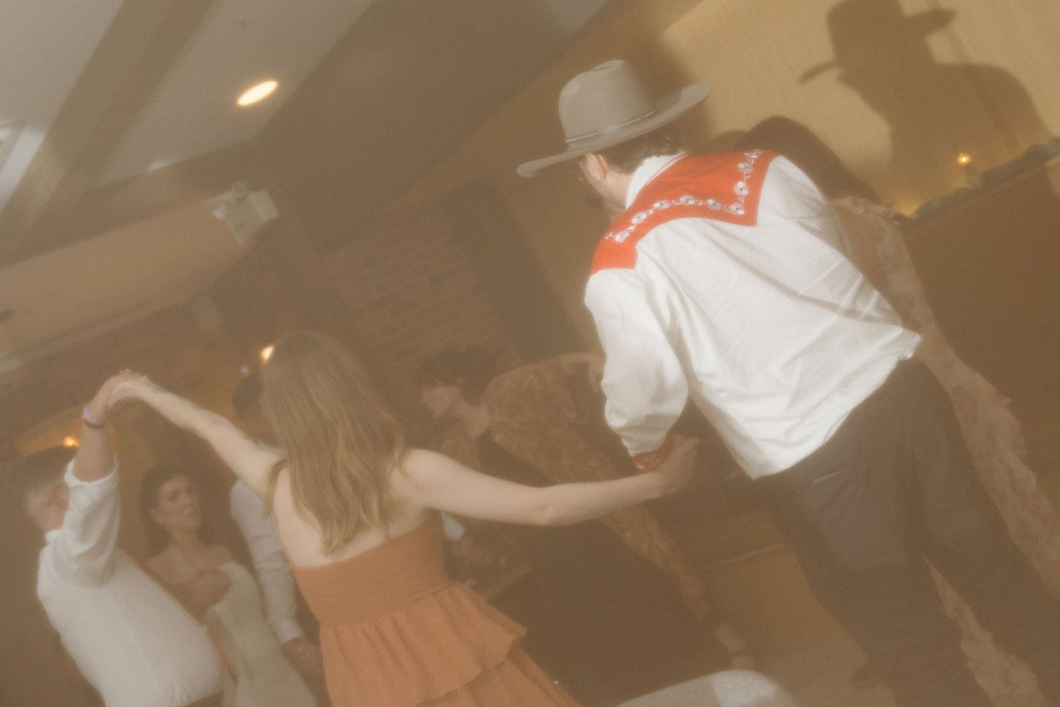 A man in a cowboy hat and white western shirt dances with a woman in an orange dress at a lively indoor party.