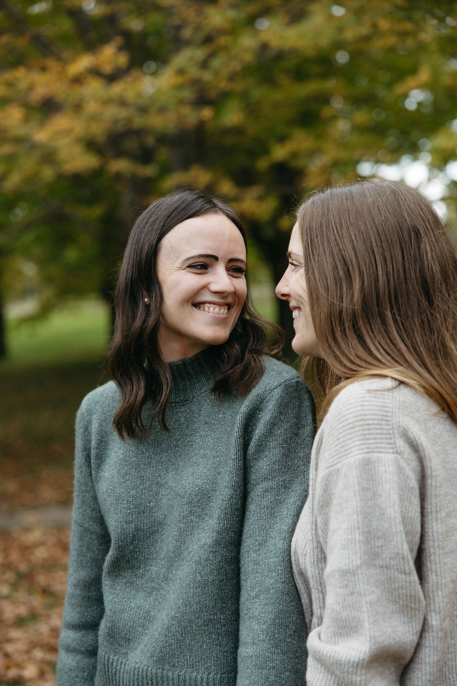 Two women in cozy knit sweaters smile warmly at each other outdoors in a park surrounded by golden autumn trees and fallen leaves.