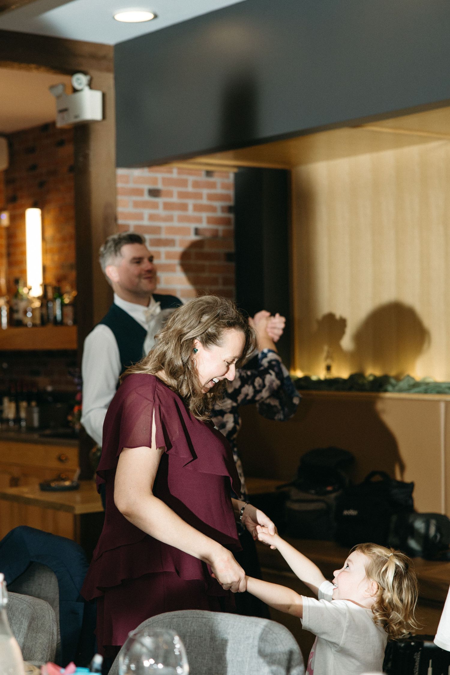 Woman in a burgundy dress dances with a smiling toddler while a man in a vest dances behind them at an indoor brick-walled venue.