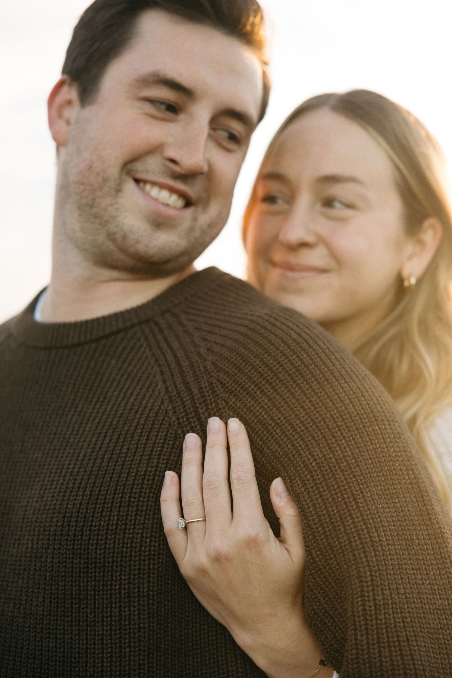 Close-up of a smiling man in a brown sweater as his partner embraces him, her engagement ring glowing in sunlight.