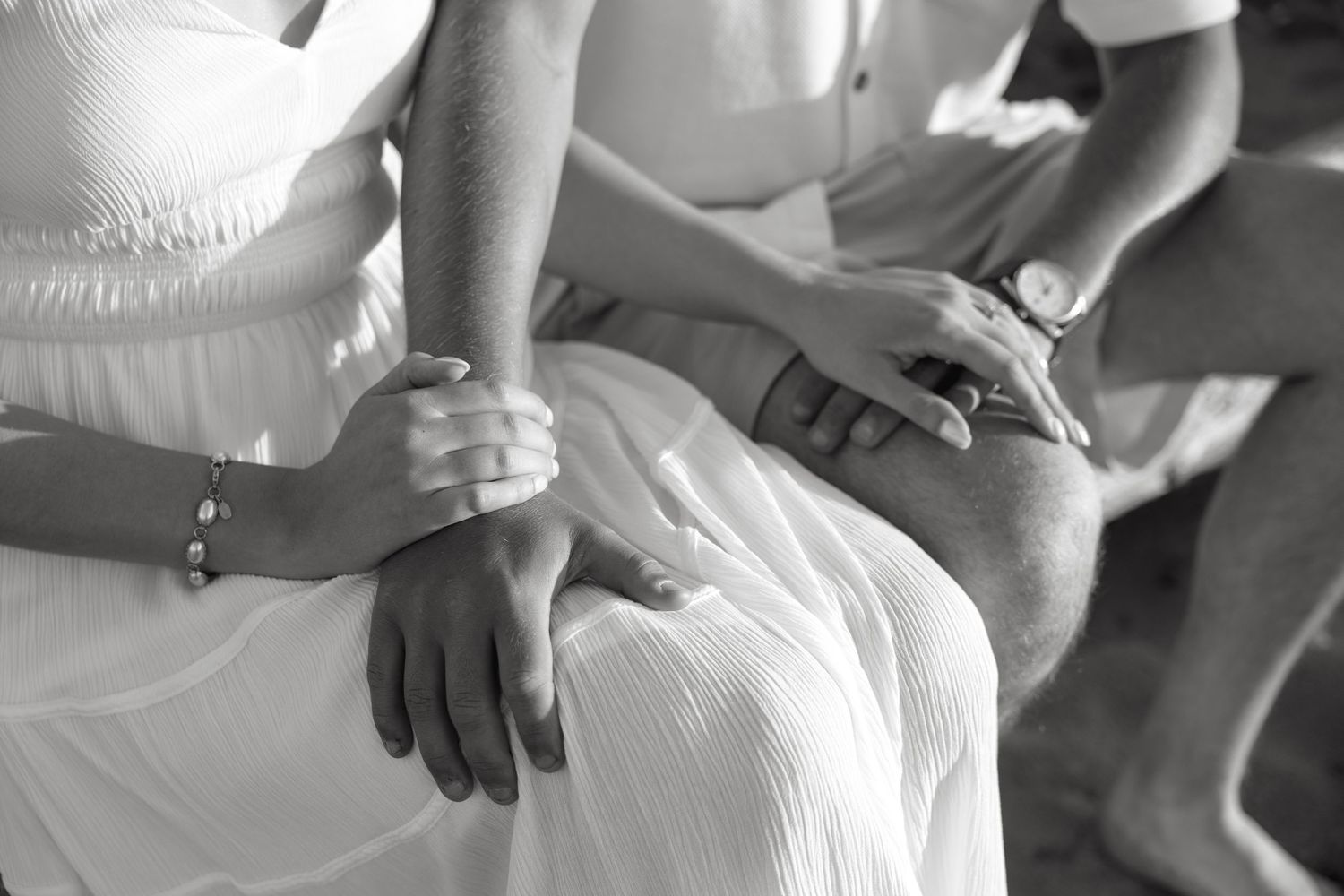 Black and white photo of a couple holding hands, wearing wedding attire, with a bracelet and watch visible.