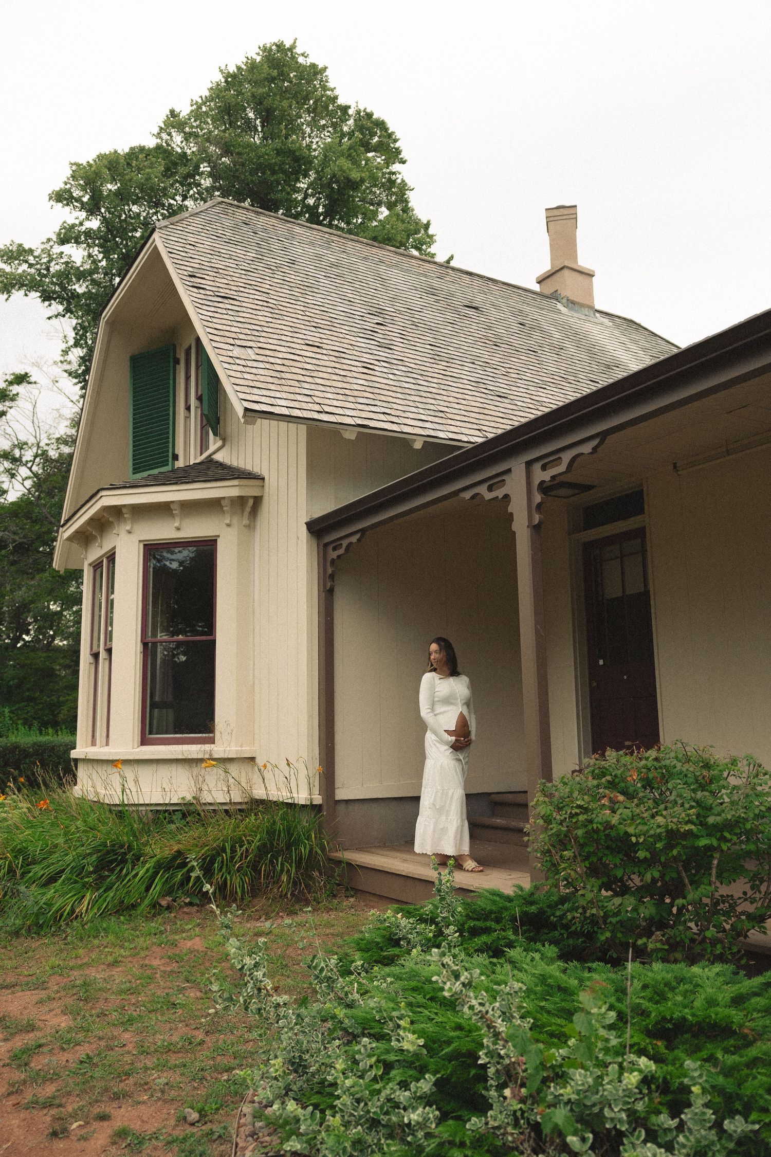 A woman in a white dress stands on the porch of a charming vintage cottage surrounded by greenery.