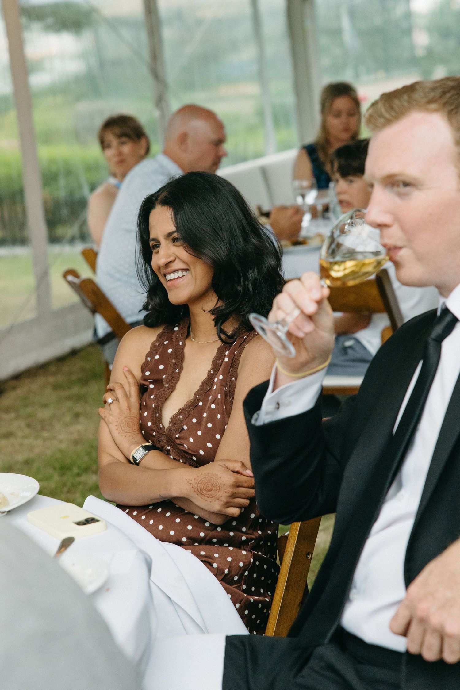 Laughing woman in brown polka dot dress sits beside a man in a tuxedo drinking wine at a wedding reception.