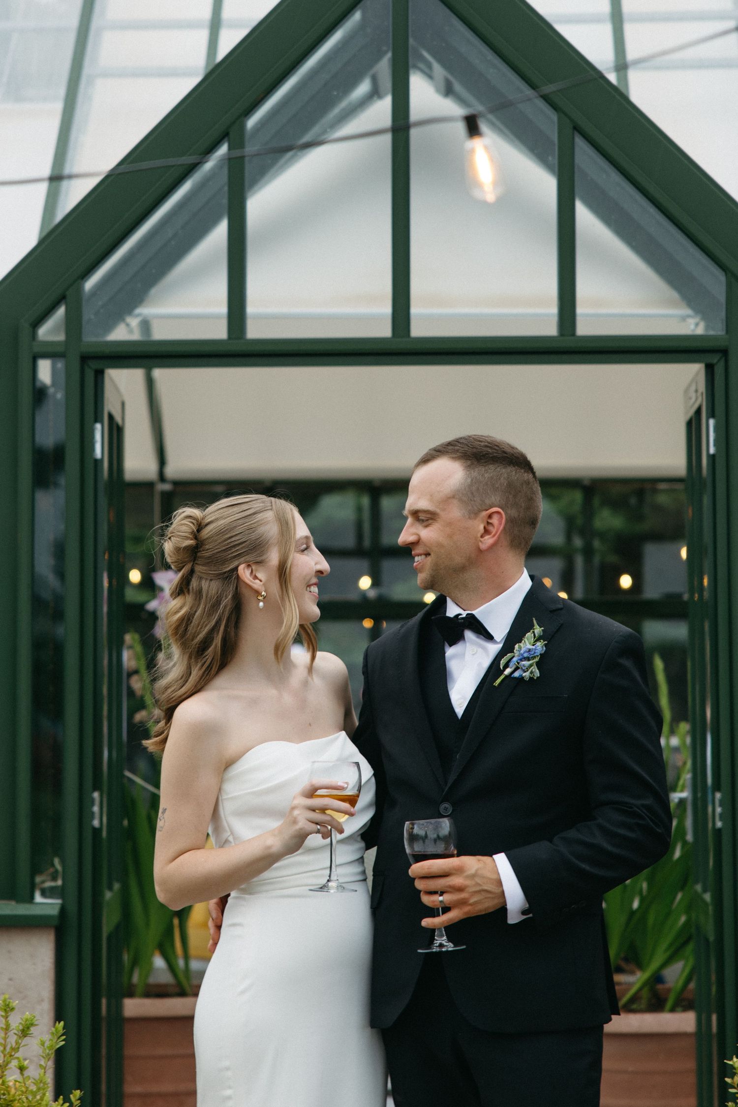 Bride and groom smiling at each other holding champagne glasses in front of a green greenhouse structure.