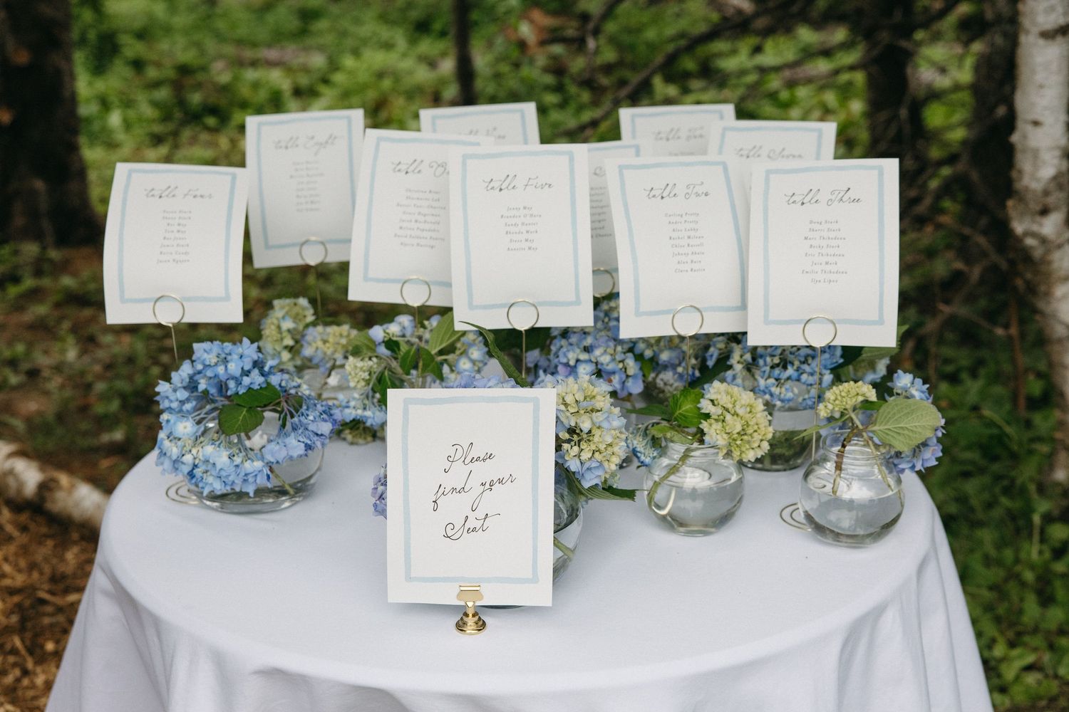 Elegant wedding seating chart display with blue hydrangea floral arrangements on a white linen-covered table.