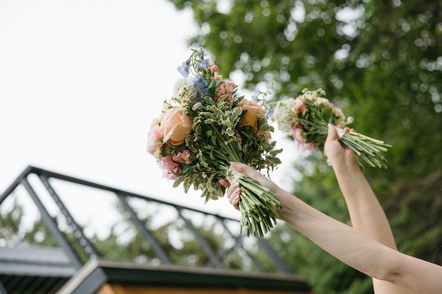 Two hands holding colorful wedding bouquets with pink roses and greenery raised against an outdoor backdrop.