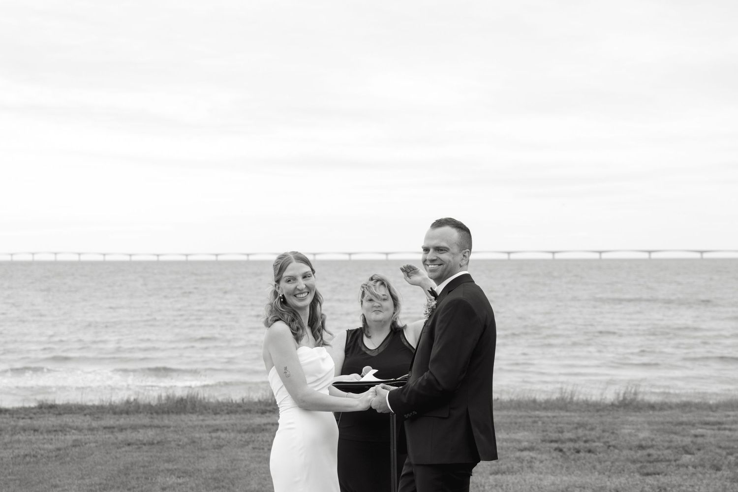Smiling bride and groom stand with officiant during outdoor waterfront wedding ceremony in black and white photo.