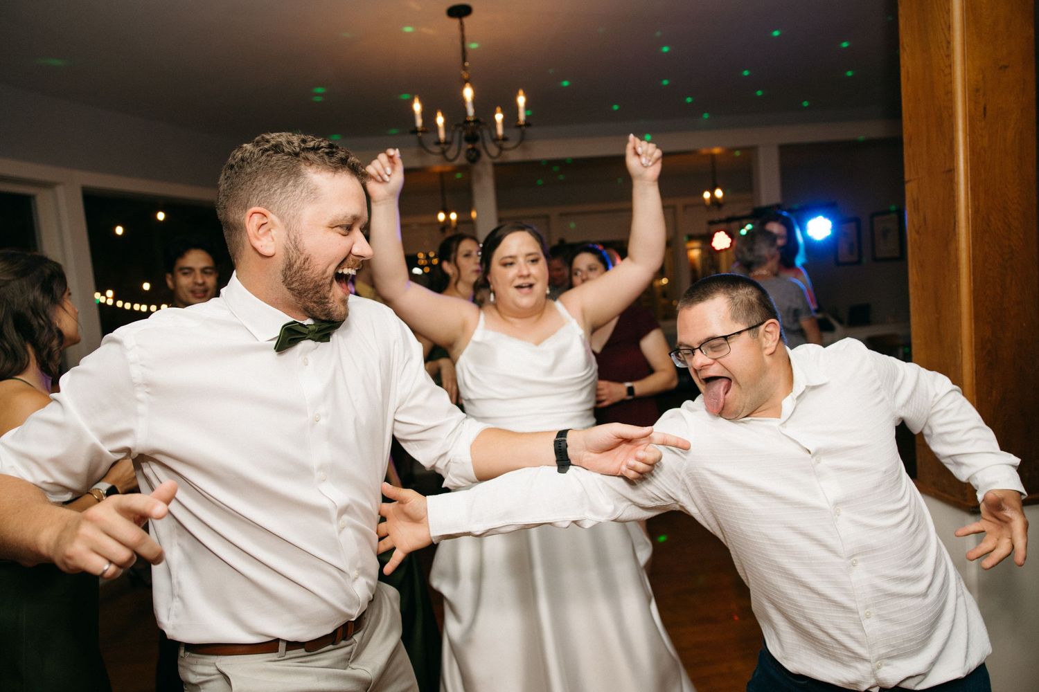 Bride and groom dancing joyfully with a guest at a lively indoor wedding reception.