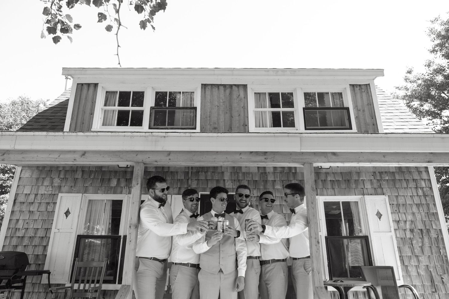 Groom and groomsmen in gray suits toasting outside a rustic shingle house in black and white photo.
