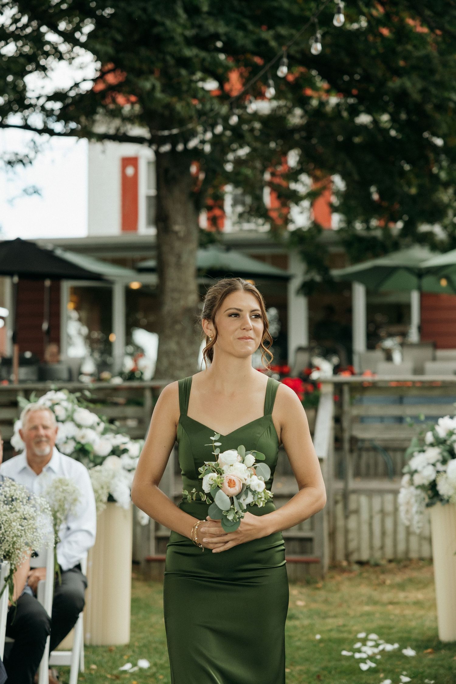 Bridesmaid in elegant green dress walks down outdoor aisle holding white floral bouquet at wedding.
