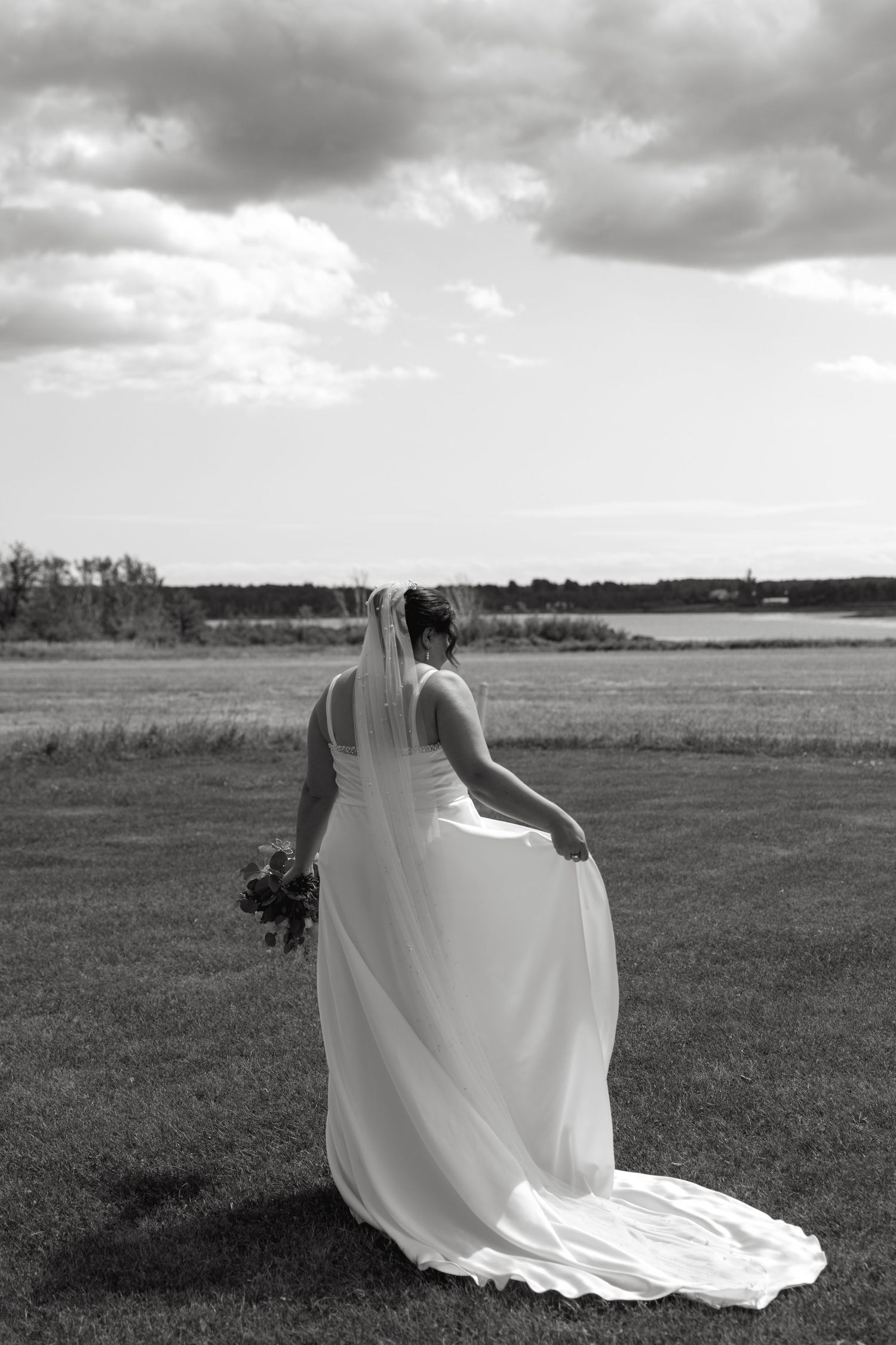 Bride in white gown and veil stands facing waterfront landscape in black and white photo.