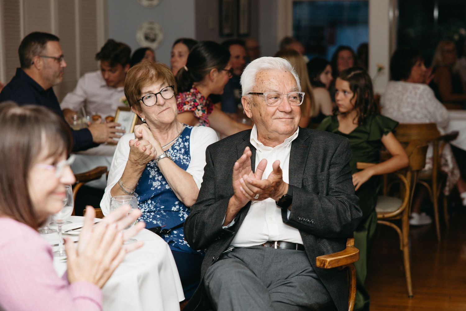 Elderly wedding guests clapping and smiling during a reception celebration in a warmly lit venue.