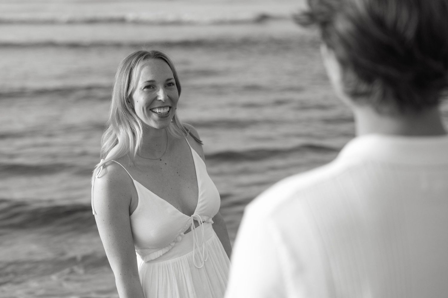 Black and white photo of a smiling blonde woman in a white dress standing on a beach.
