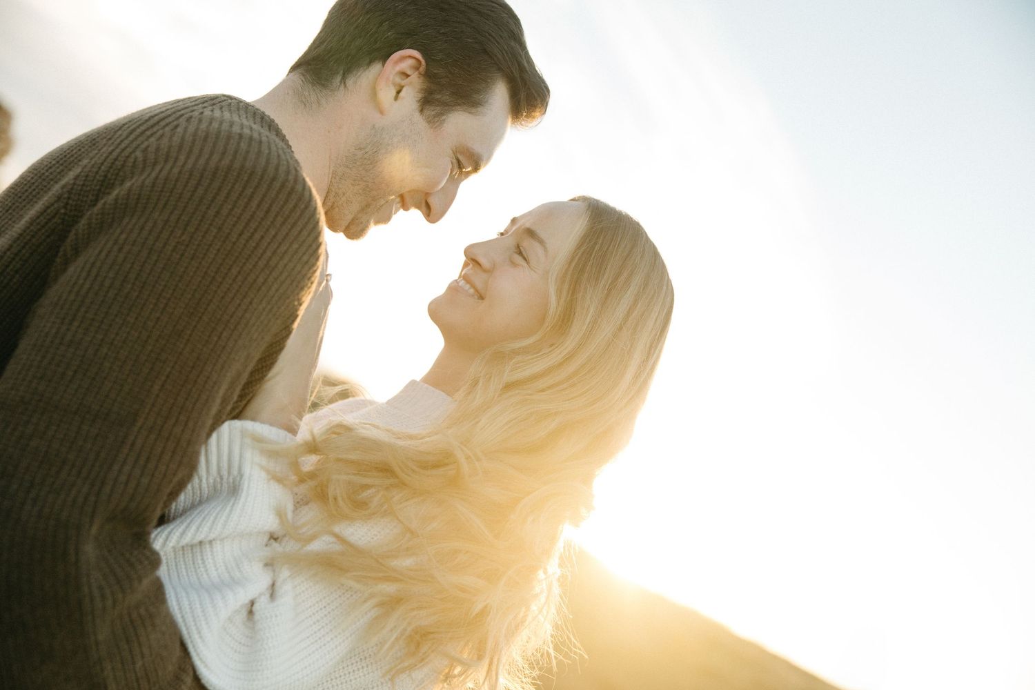 Smiling couple embracing outdoors in warm golden sunlight as they gaze lovingly at each other, the woman with long wavy blonde hair.