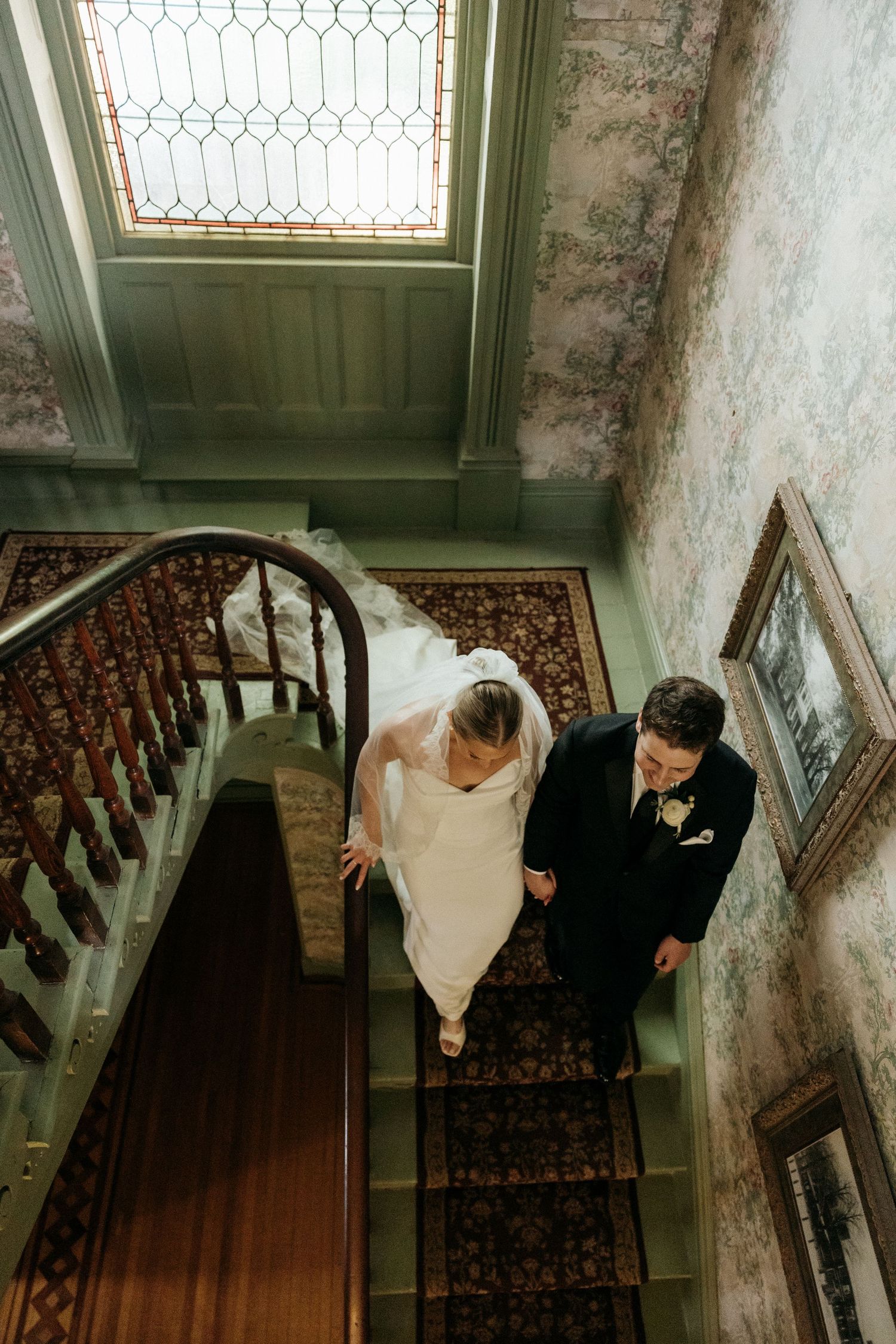 A couple descends an ornate vintage staircase with stained glass windows during their wedding.