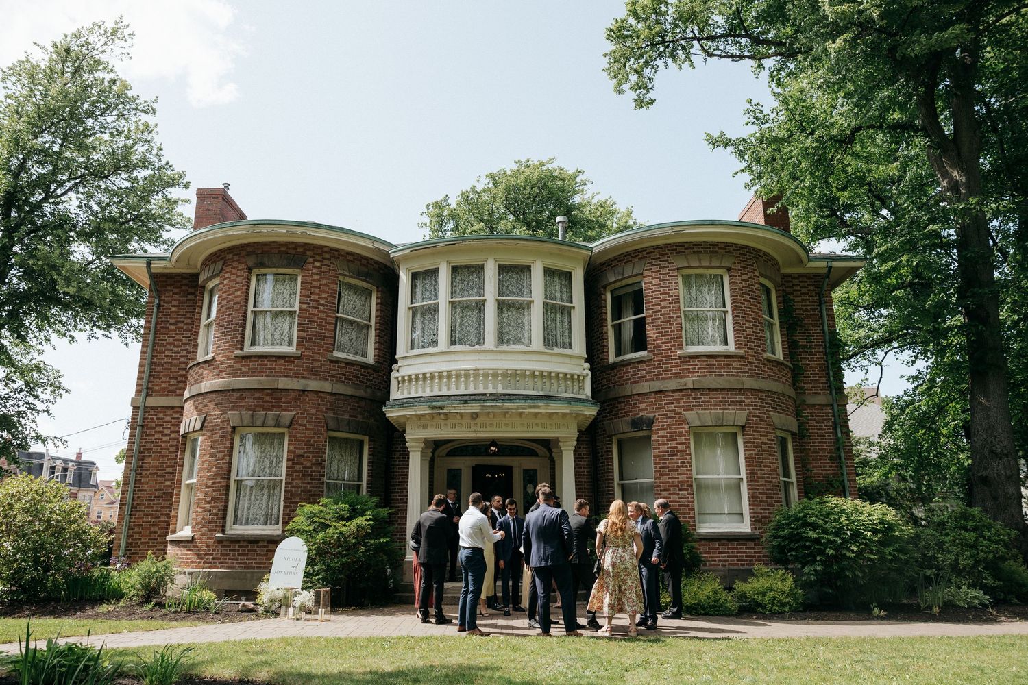 Panoramic view of a historic brick mansion with guests gathered for an outdoor wedding ceremony.
