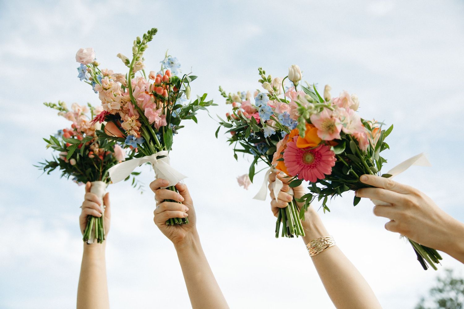 Hands raised holding colorful wedding bouquets against blue sky.