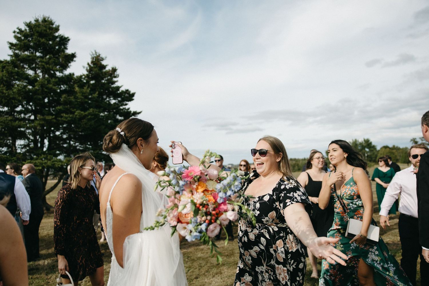 Wedding guests gather on a grassy field with evergreen trees in the background.