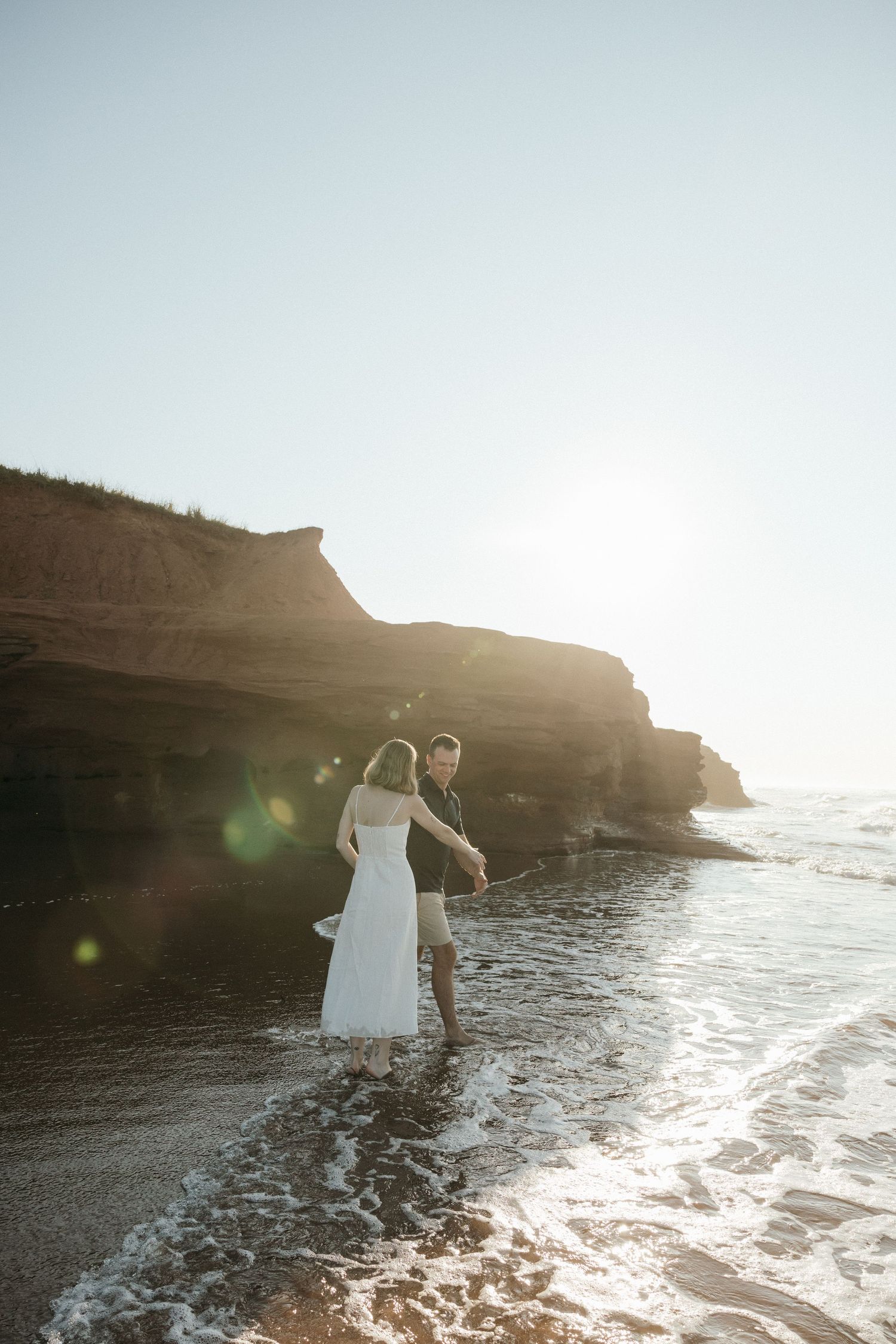 A couple stands in ocean waves near dramatic coastal cliffs at sunset.