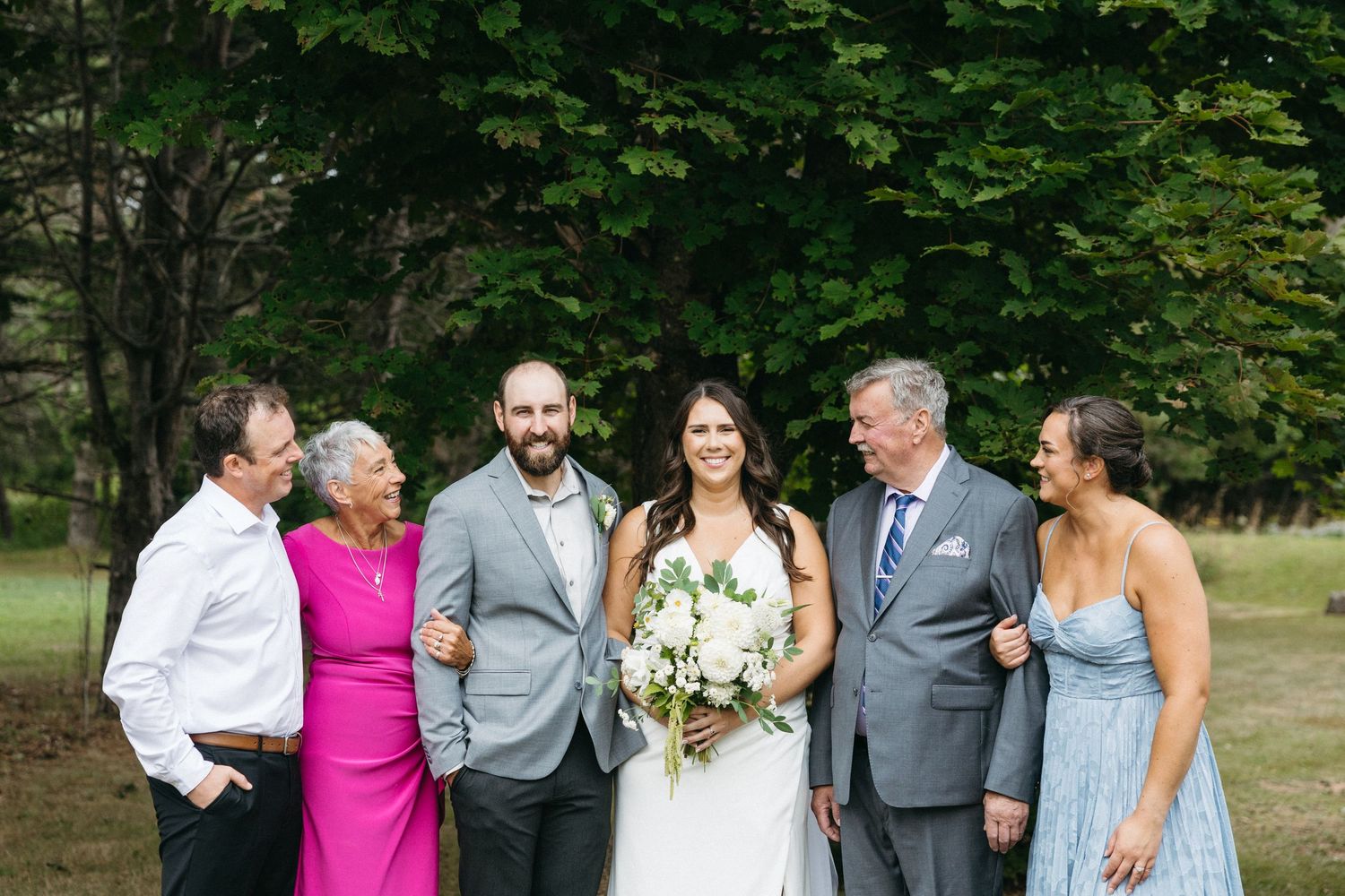Wedding party members pose for casual photos on grass with trees in background.