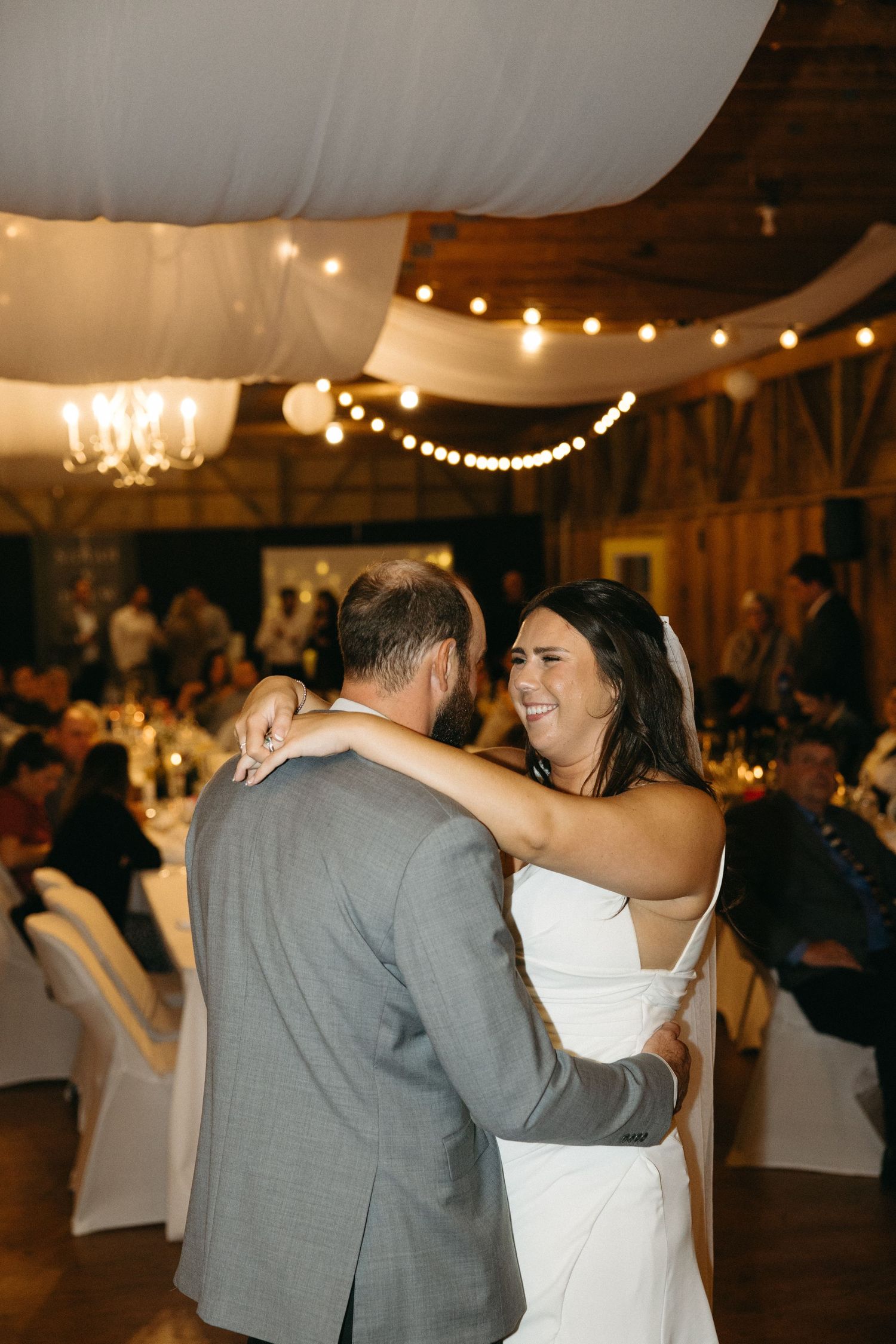 Couple shares romantic dance under string lights at rustic barn wedding reception.