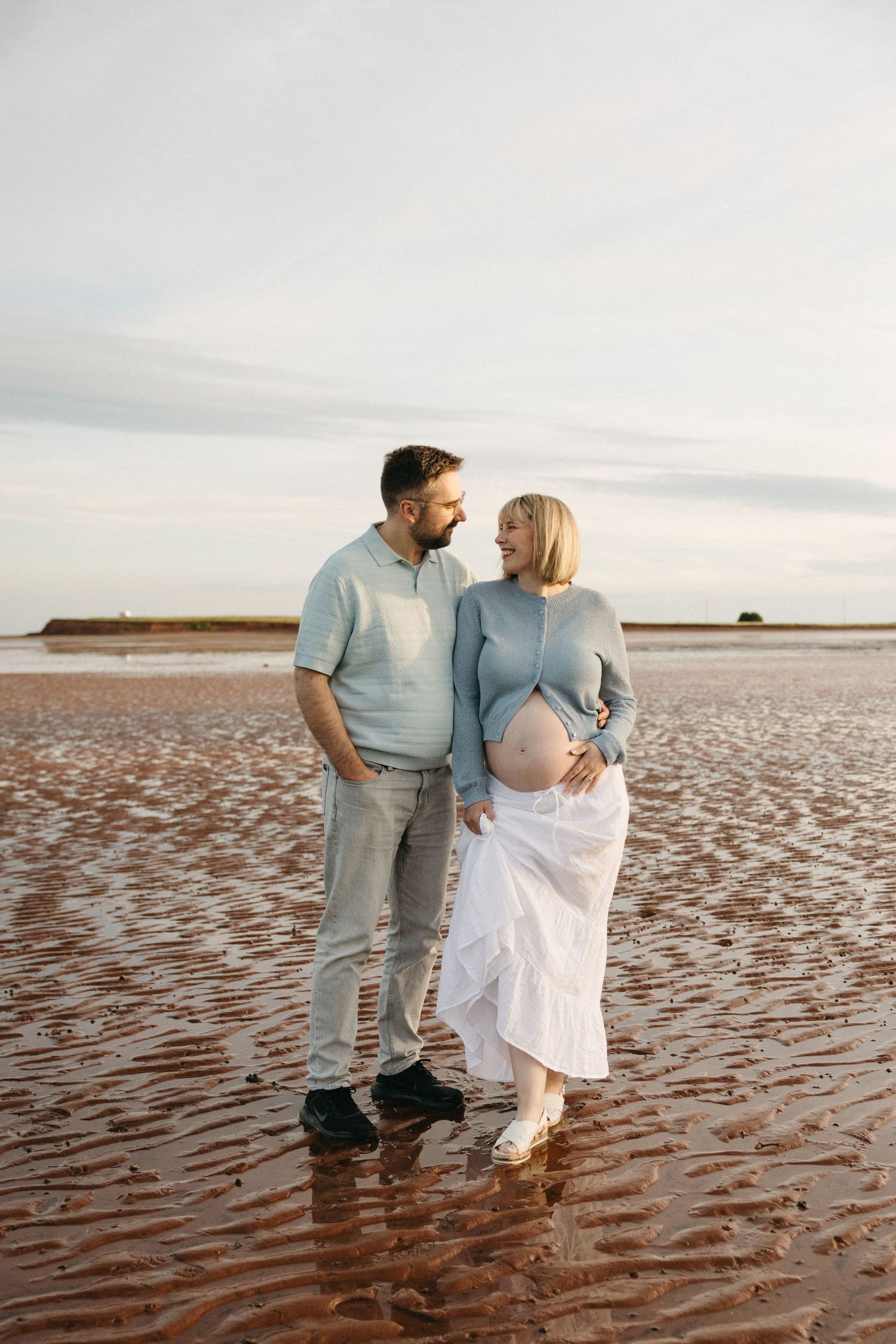 A pregnant couple stands on a wet sandy beach, smiling at each other, the woman cradling her baby bump in a white skirt.