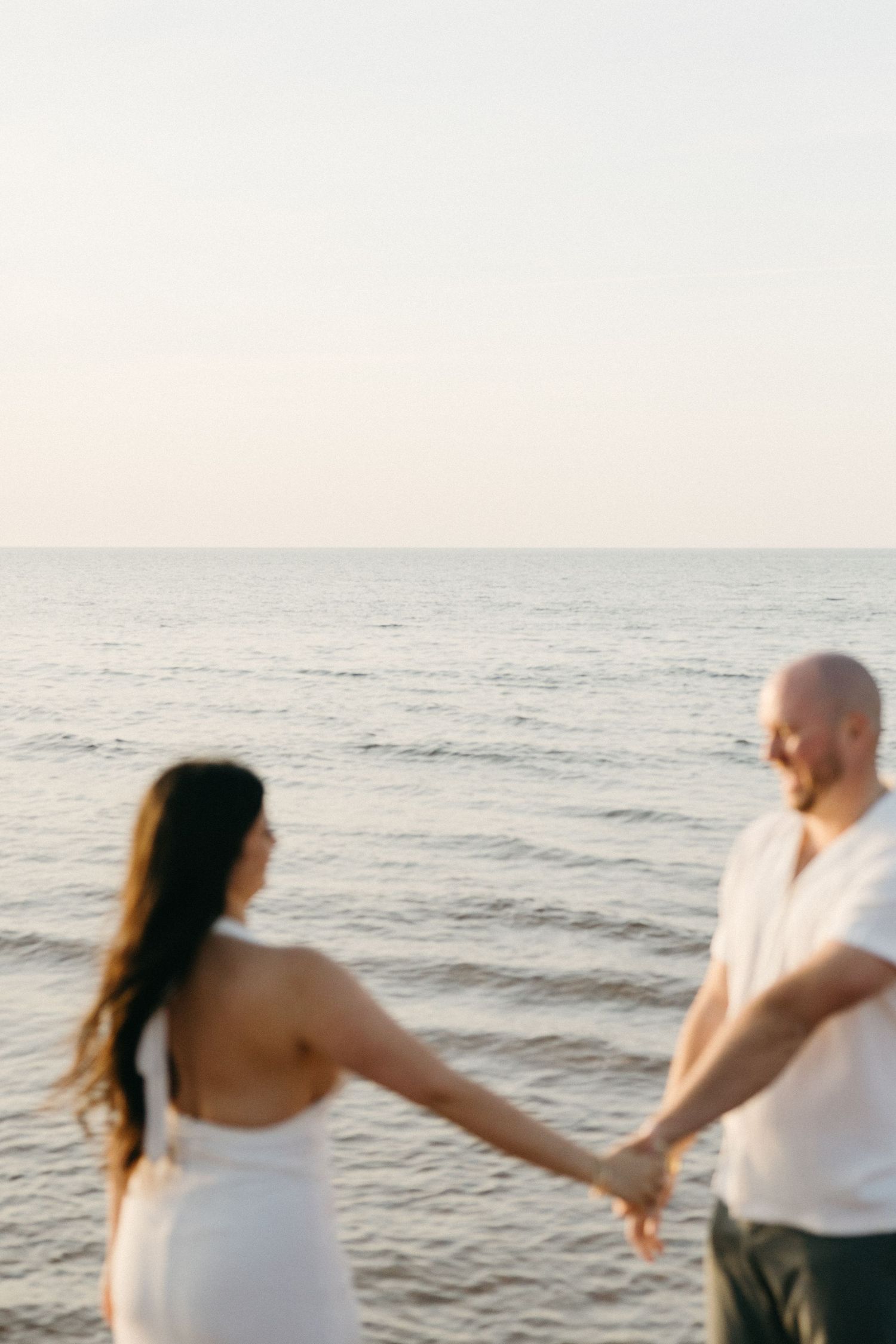 Couple in white outfits holding hands facing the calm ocean at sunset on the beach.