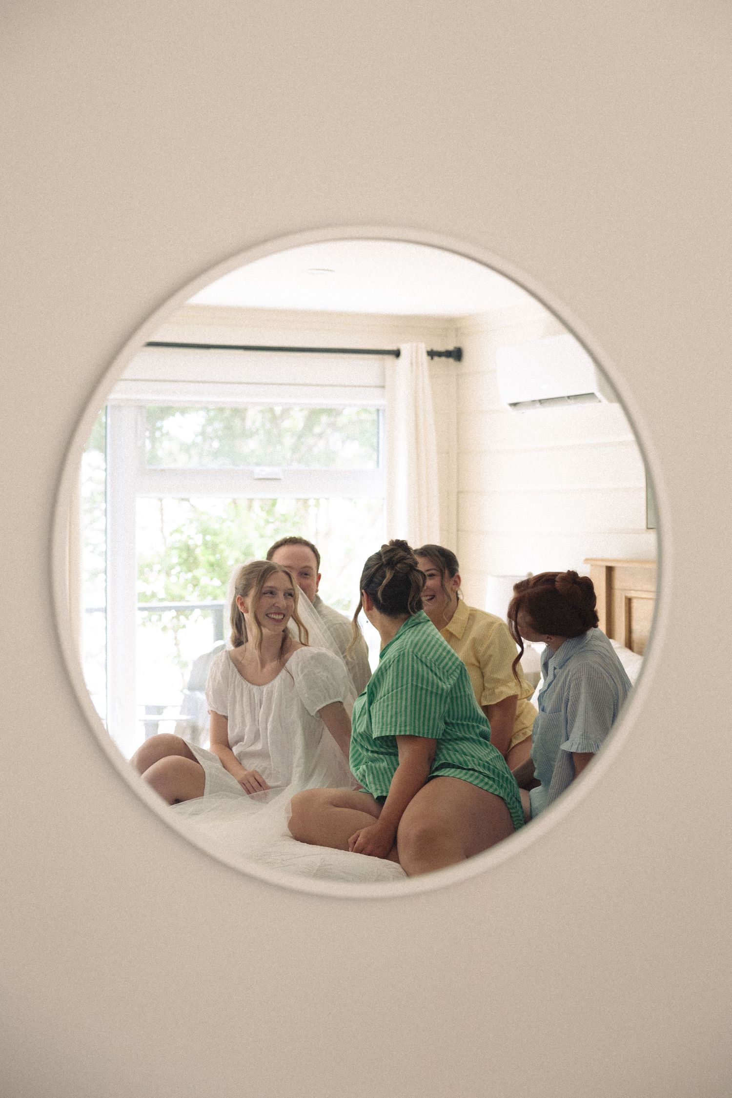 Group of women gathered in a bright bedroom, viewed through a round porthole window opening in a white wall.