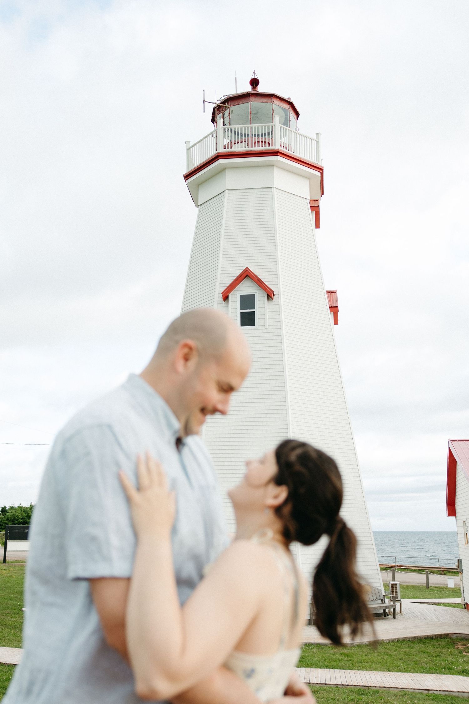 A couple shares a romantic moment in front of a white and red lighthouse by the sea.