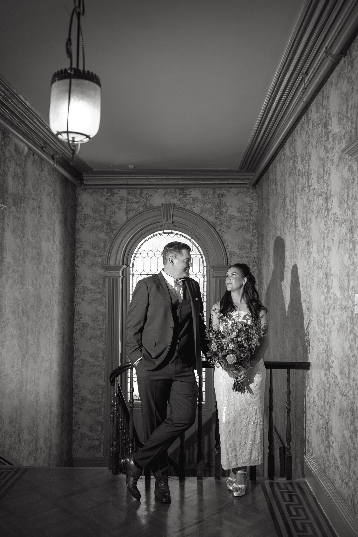 Black and white photo of a groom and bride holding a bouquet, standing in a grand hallway beneath an arched window.