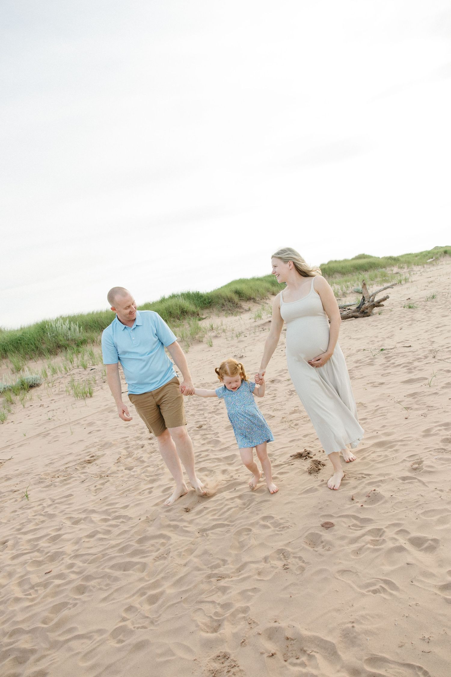 A happy family walks barefoot on sandy beach dunes, with a pregnant woman in a white dress holding hands with a toddler.