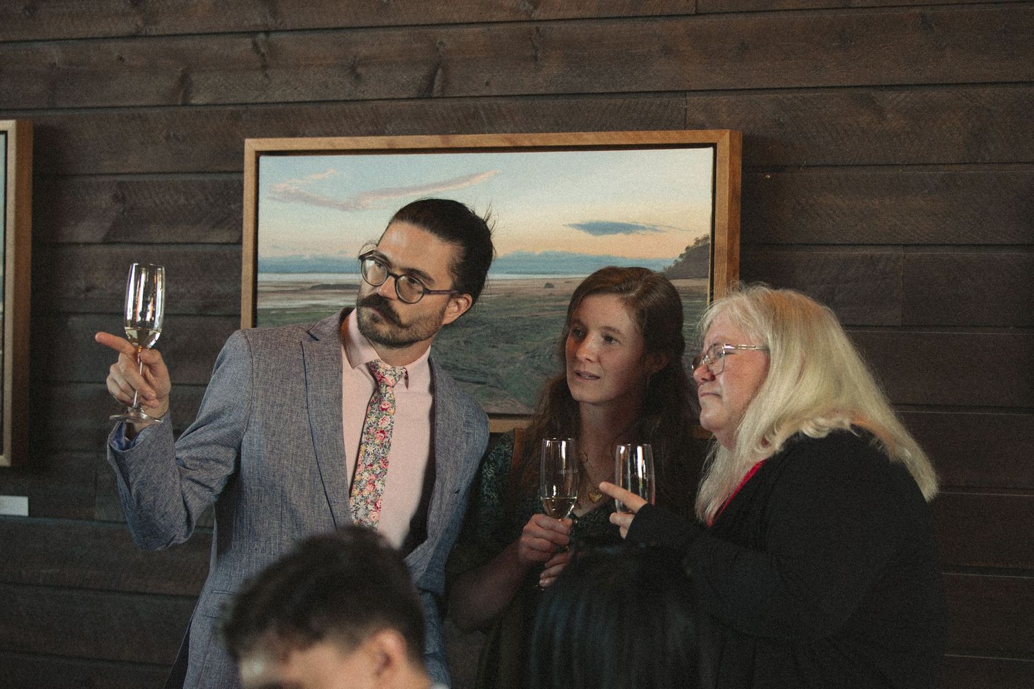 Wedding guests enjoying drinks and conversation against a rustic wooden wall with framed artwork.