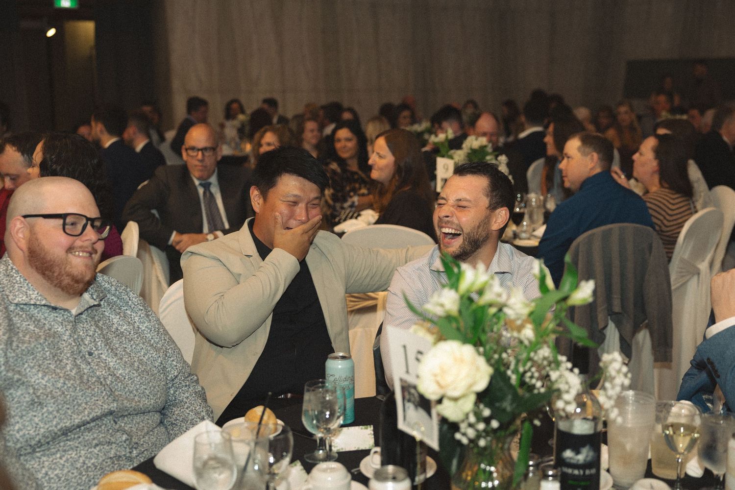 Wedding guests sharing laughter at a reception dinner table with white floral decorations.