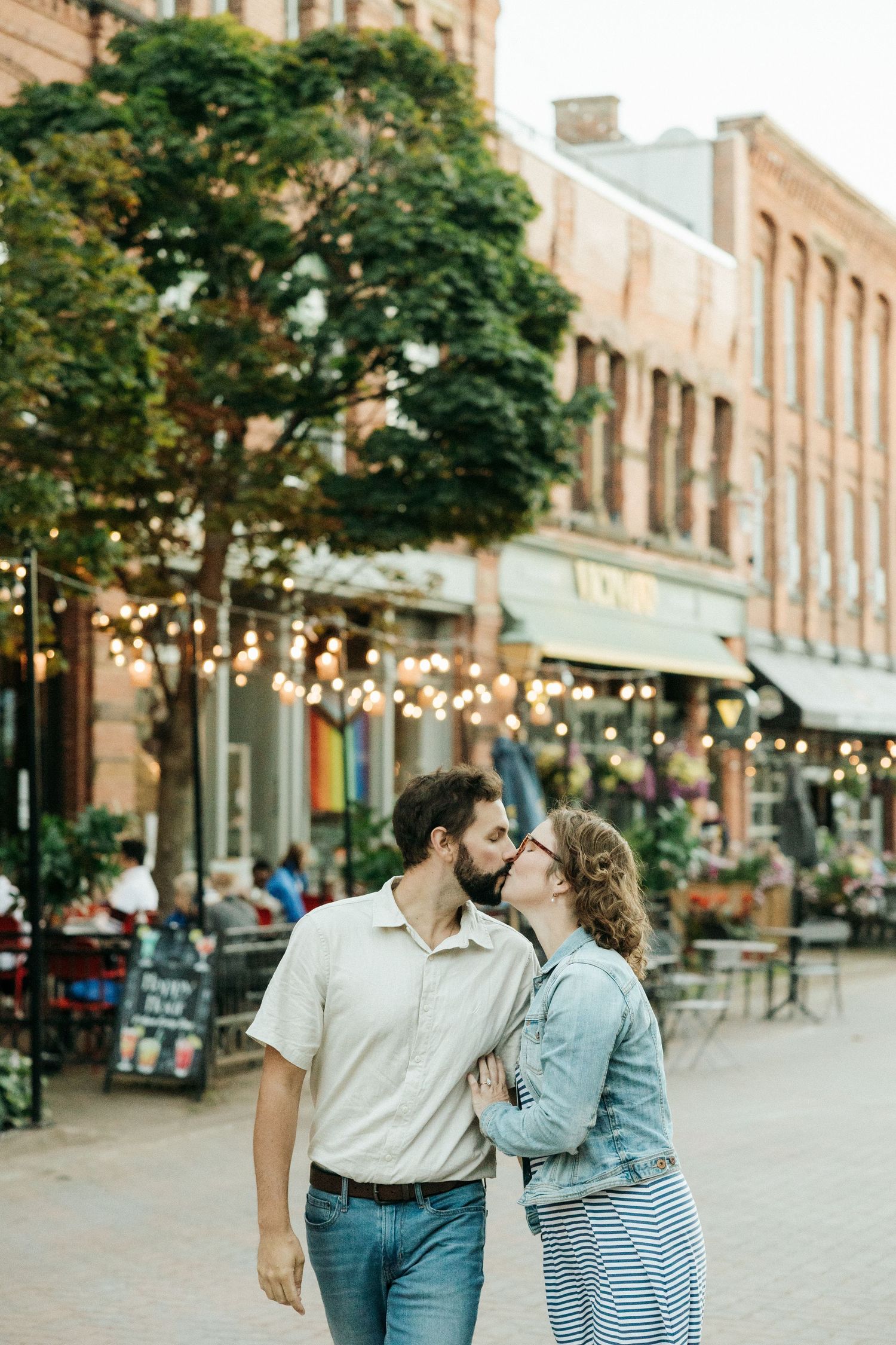 A couple shares an intimate moment on a downtown street with twinkling lights and cafes.