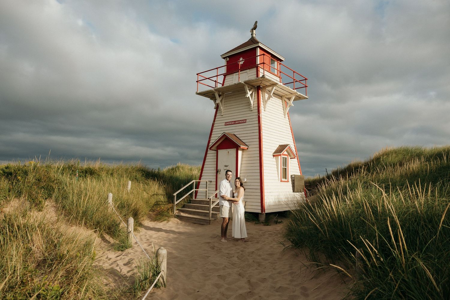 Red and white lighthouse stands tall against cloudy sky on beach path.