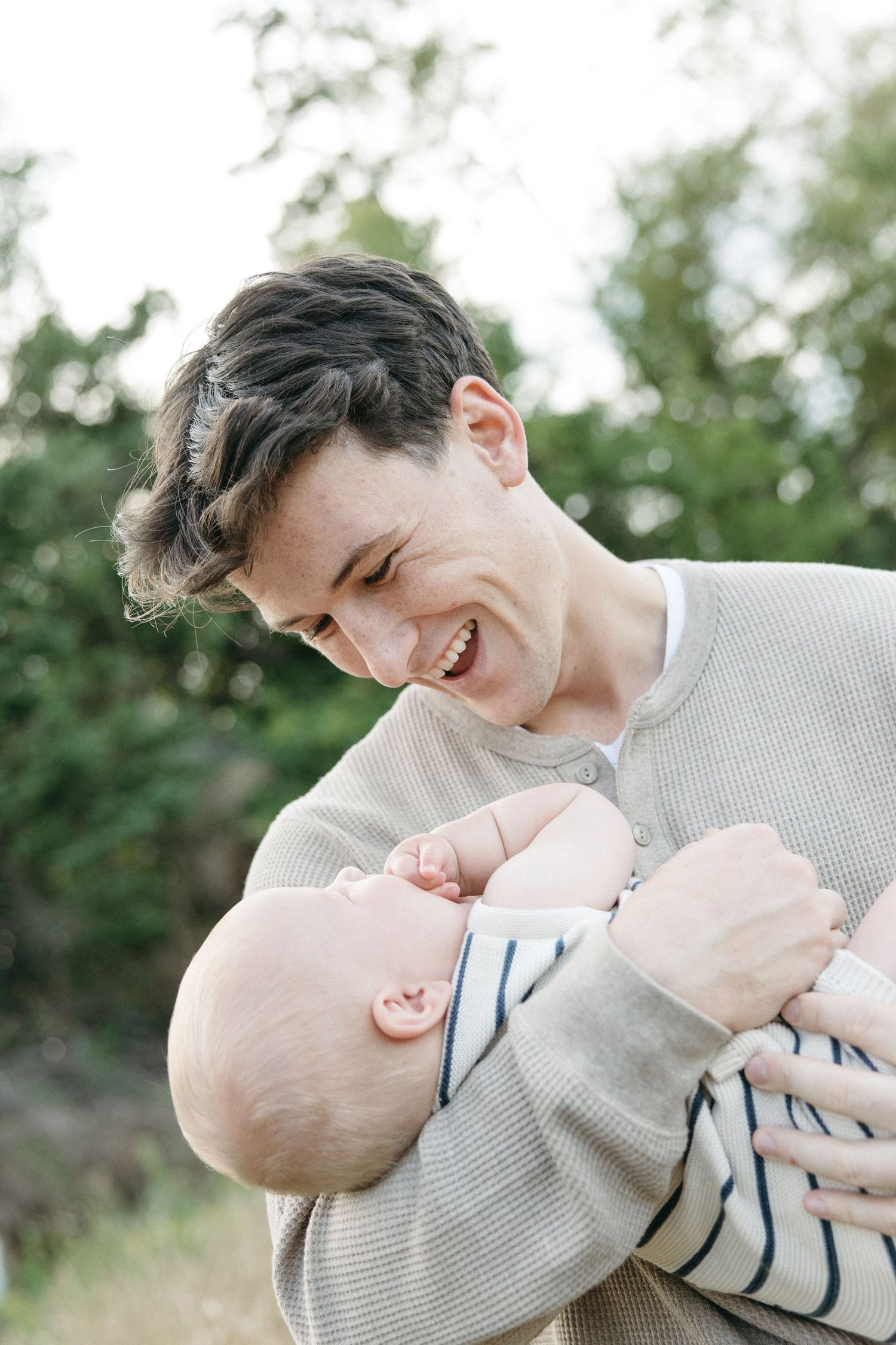 A smiling young man laughs while cradling a happy baby outdoors among green trees.